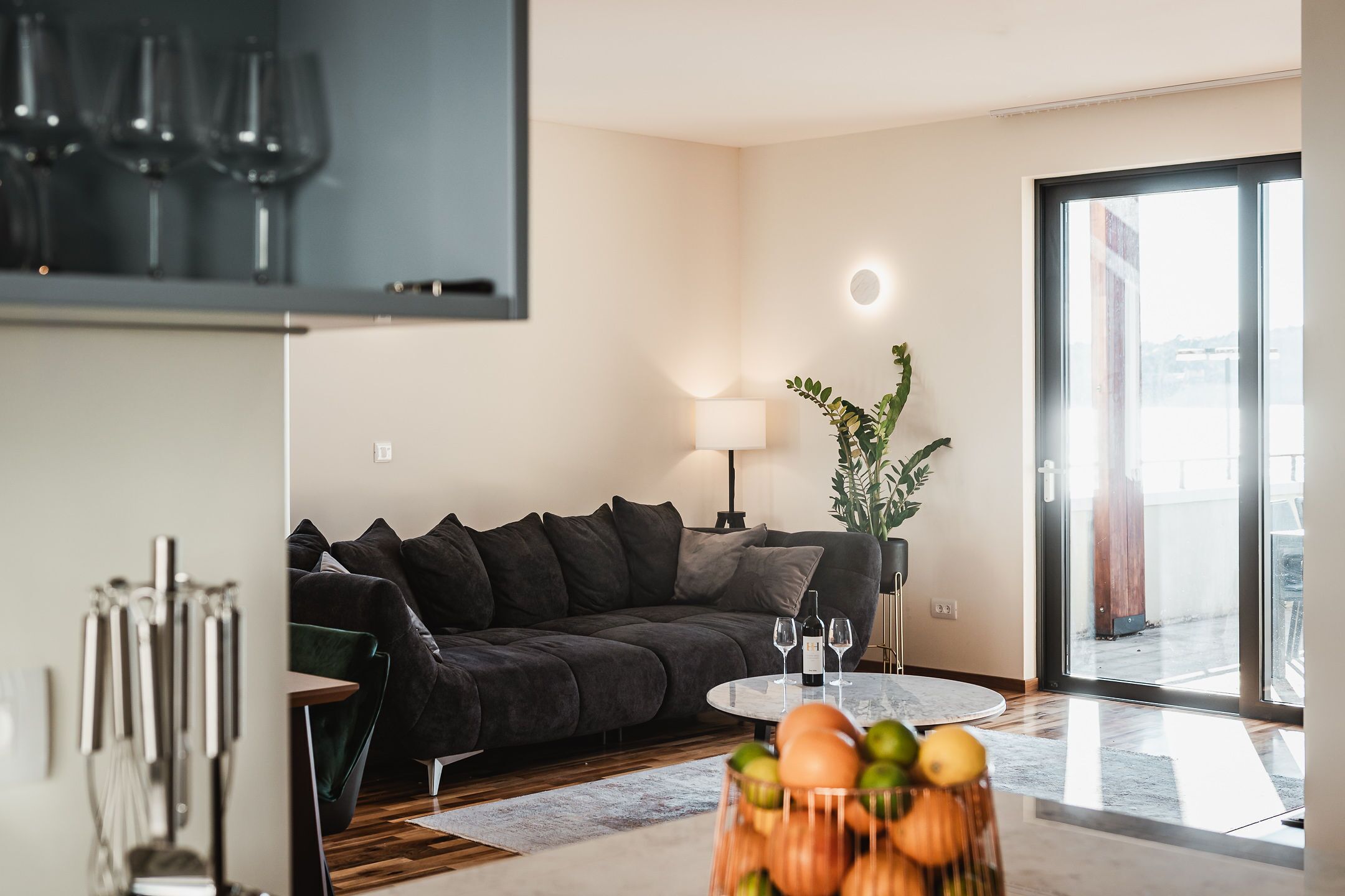 A modern living room with a dark grey sofa, a round coffee table with fruits, a TV mounted on the wall, and a large window with a view of the outside.