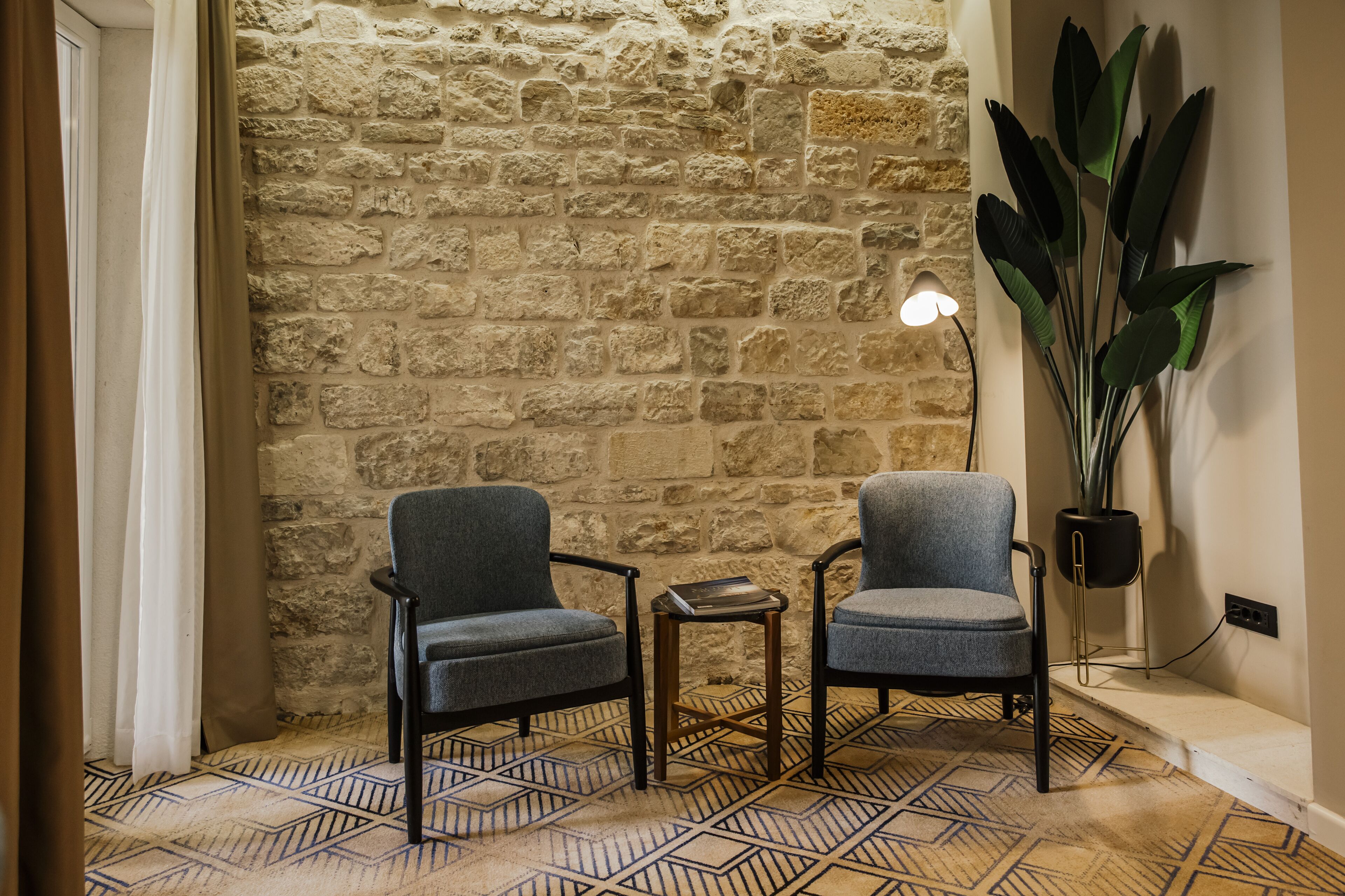 A room with two grey armchairs, a small table, a lamp, and a large plant against a stone wall.