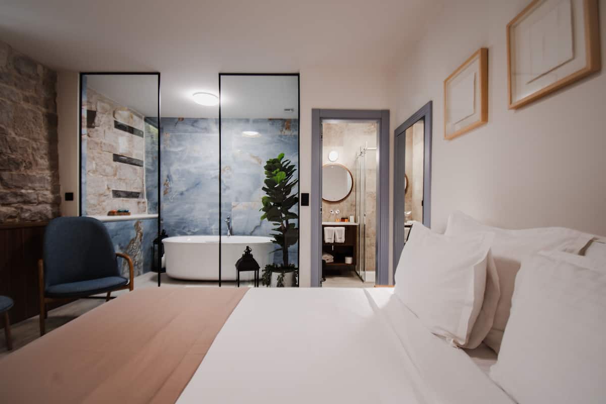 Une chambre à coucher moderne avec un grand lit, une chaise, une baignoire et une salle de bain ouverte, équipée d'un miroir et d'un porte-serviettes.