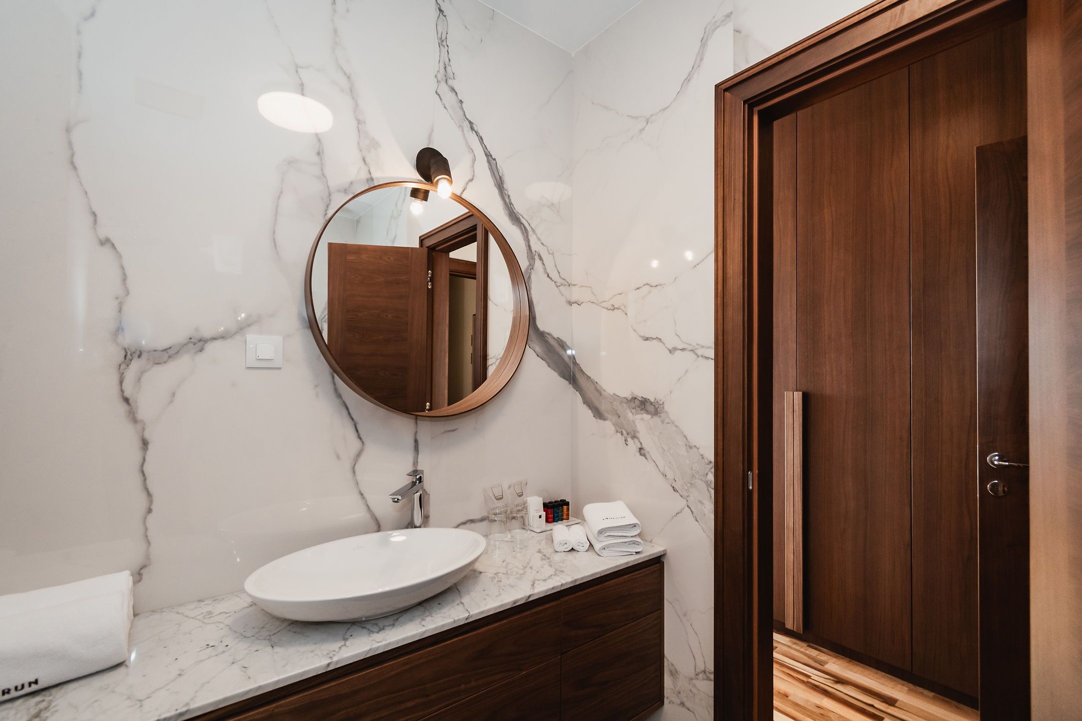 A bathroom with a marble wall, a round mirror, a wooden vanity, and a wooden door.