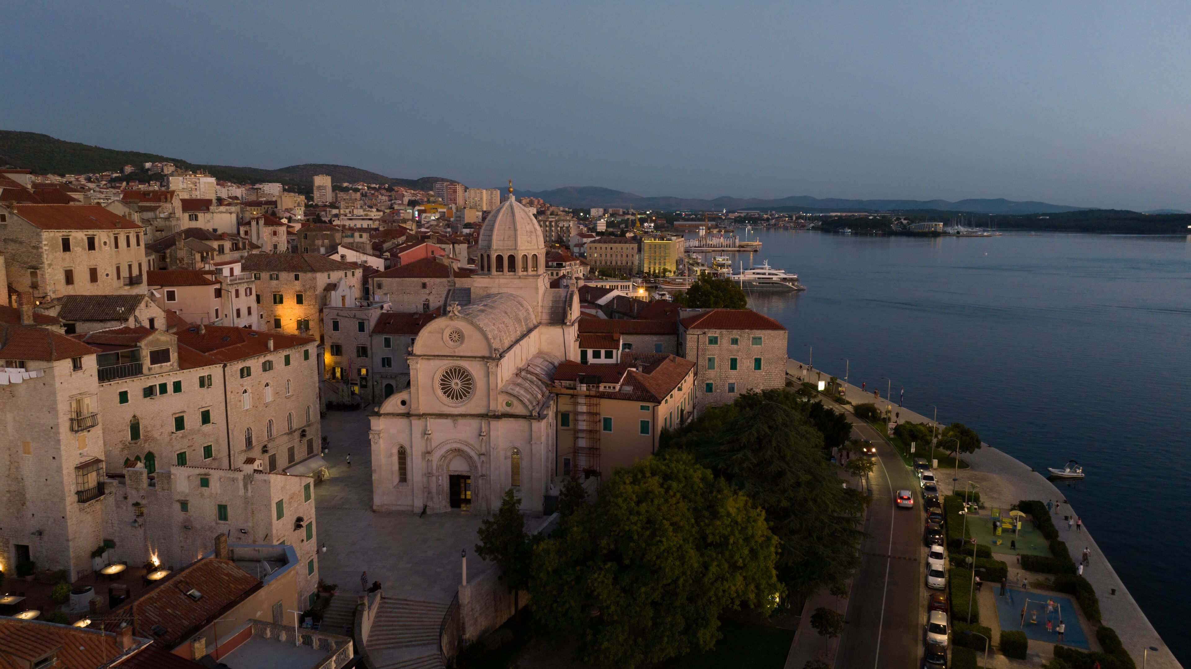 A coastal cityscape with historic architecture, including a prominent church with a dome and bell tower, situated by a waterfront with a promenade and parked cars.