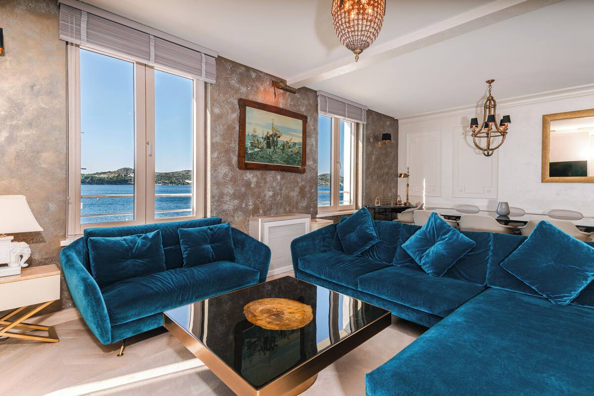 Un salon moderne avec deux canapés bleus, une table basse en verre et une vue sur la mer grâce à de grandes fenêtres.