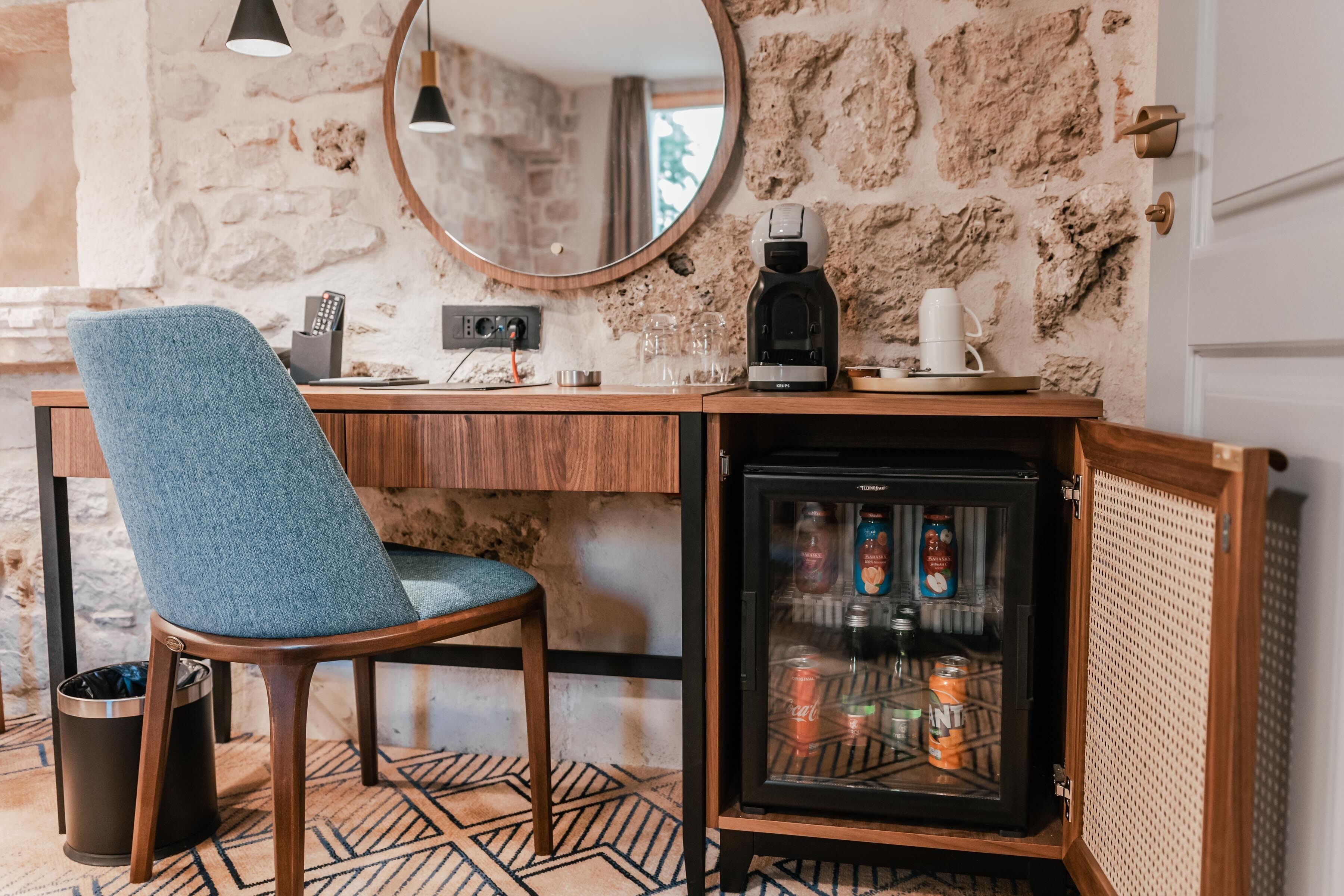 A compact kitchenette with a coffee maker, a refrigerator stocked with beverages, and a chair.