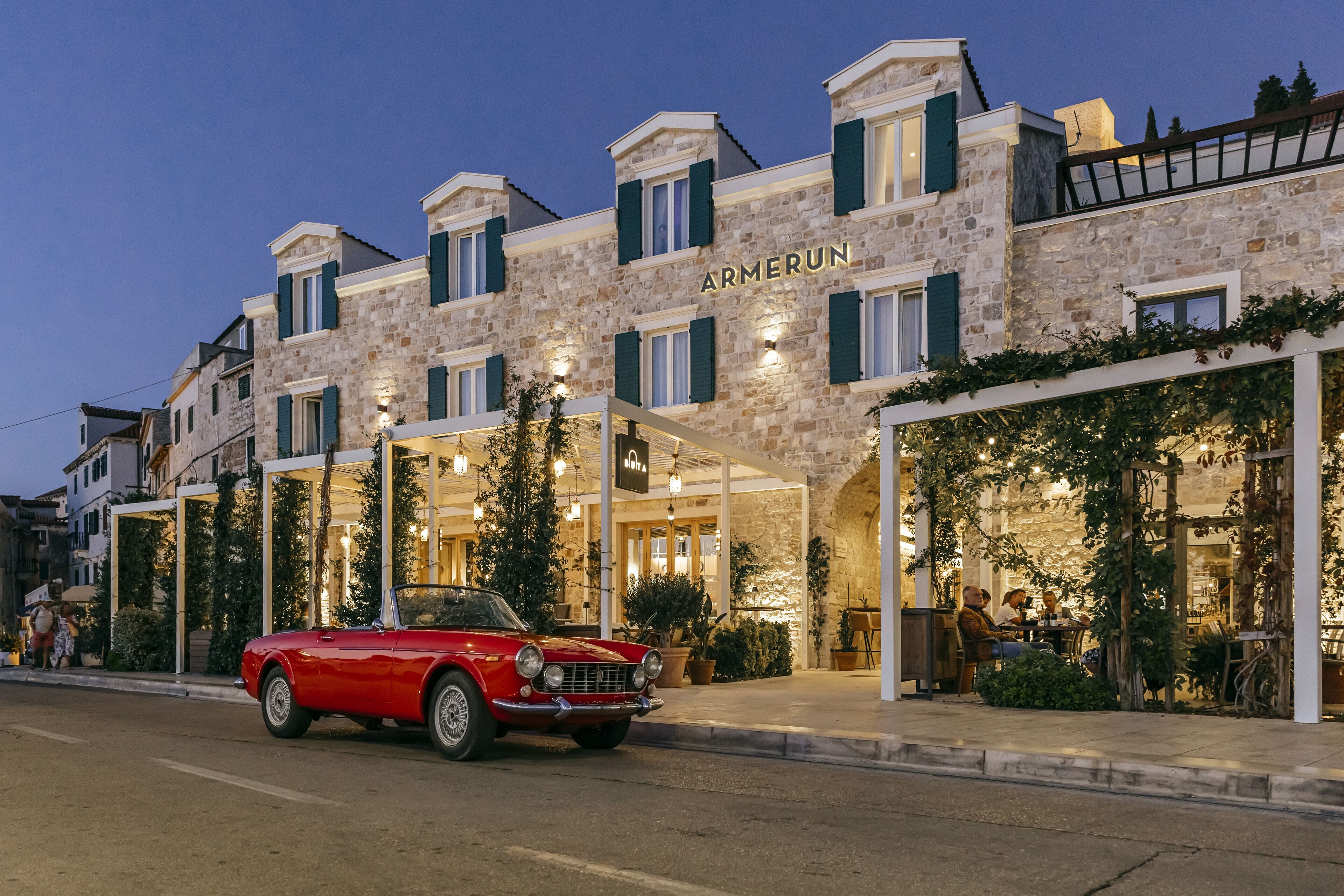 A stone building with the name "ARMERUN" on the facade, featuring a classic red convertible parked in front.