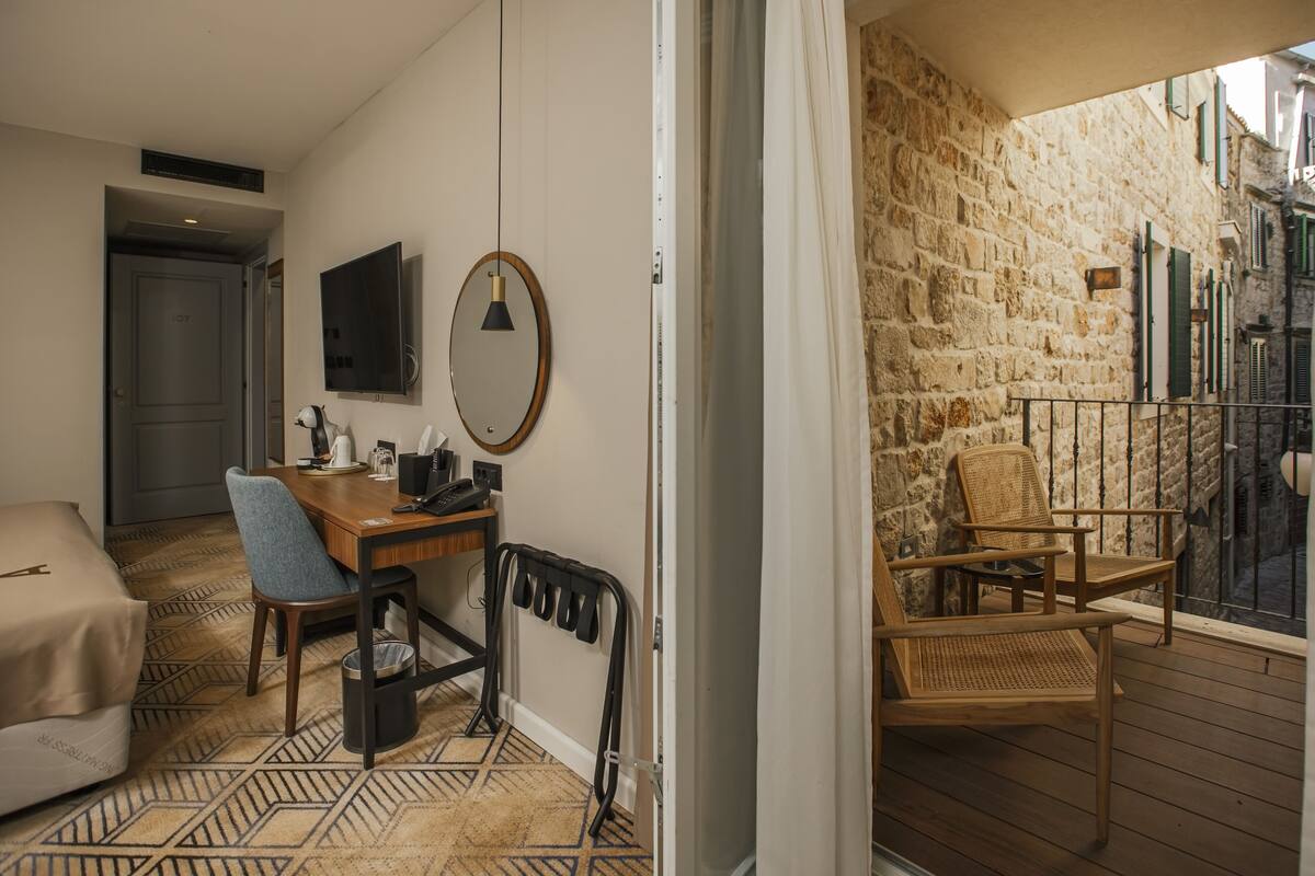 Une chambre d’hôtel avec un lit, un bureau et une chaise, un miroir et une vue sur un bâtiment en pierre à l’extérieur.