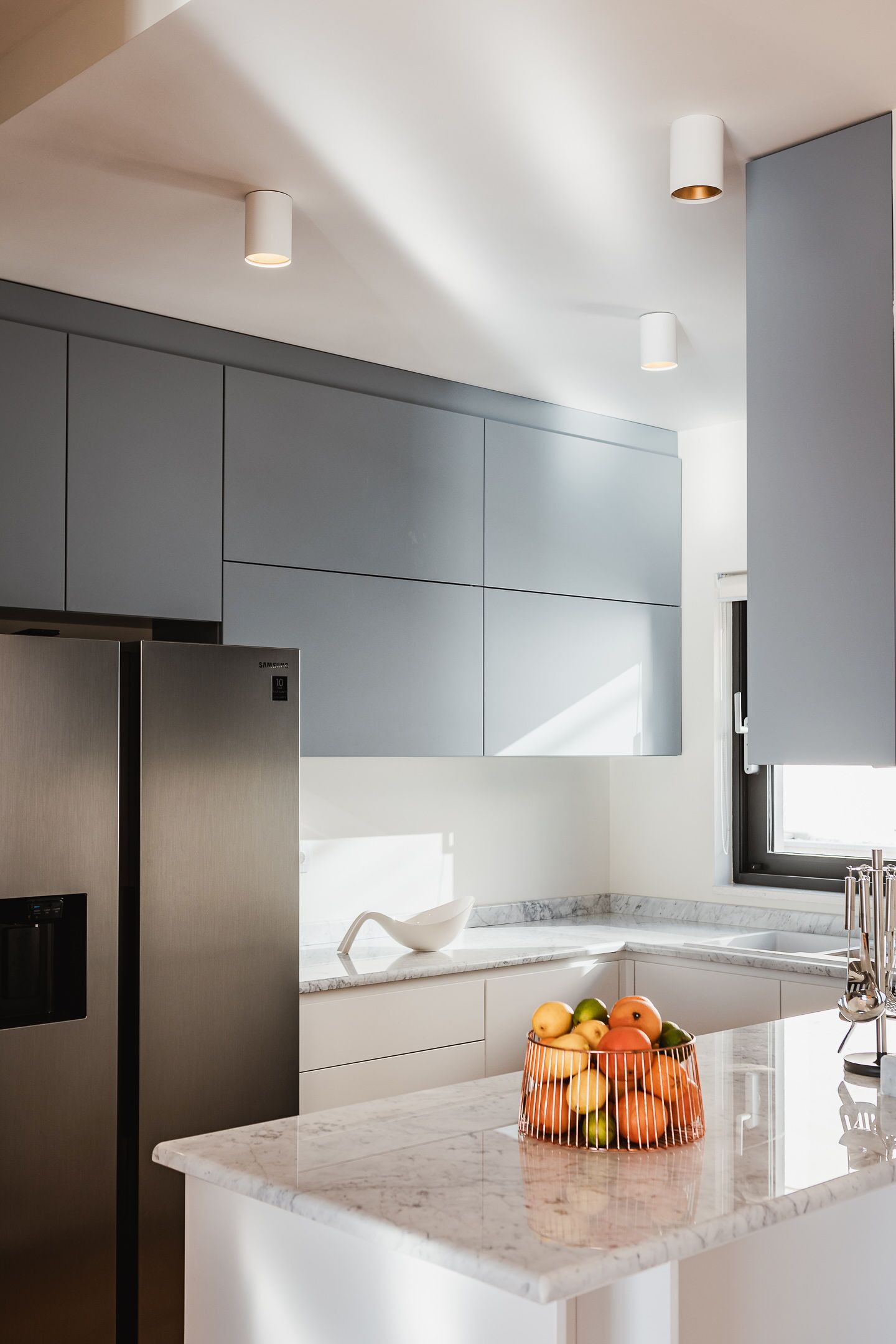 A modern kitchen with grey cabinets, a stainless steel refrigerator, and a marble countertop with a fruit basket.