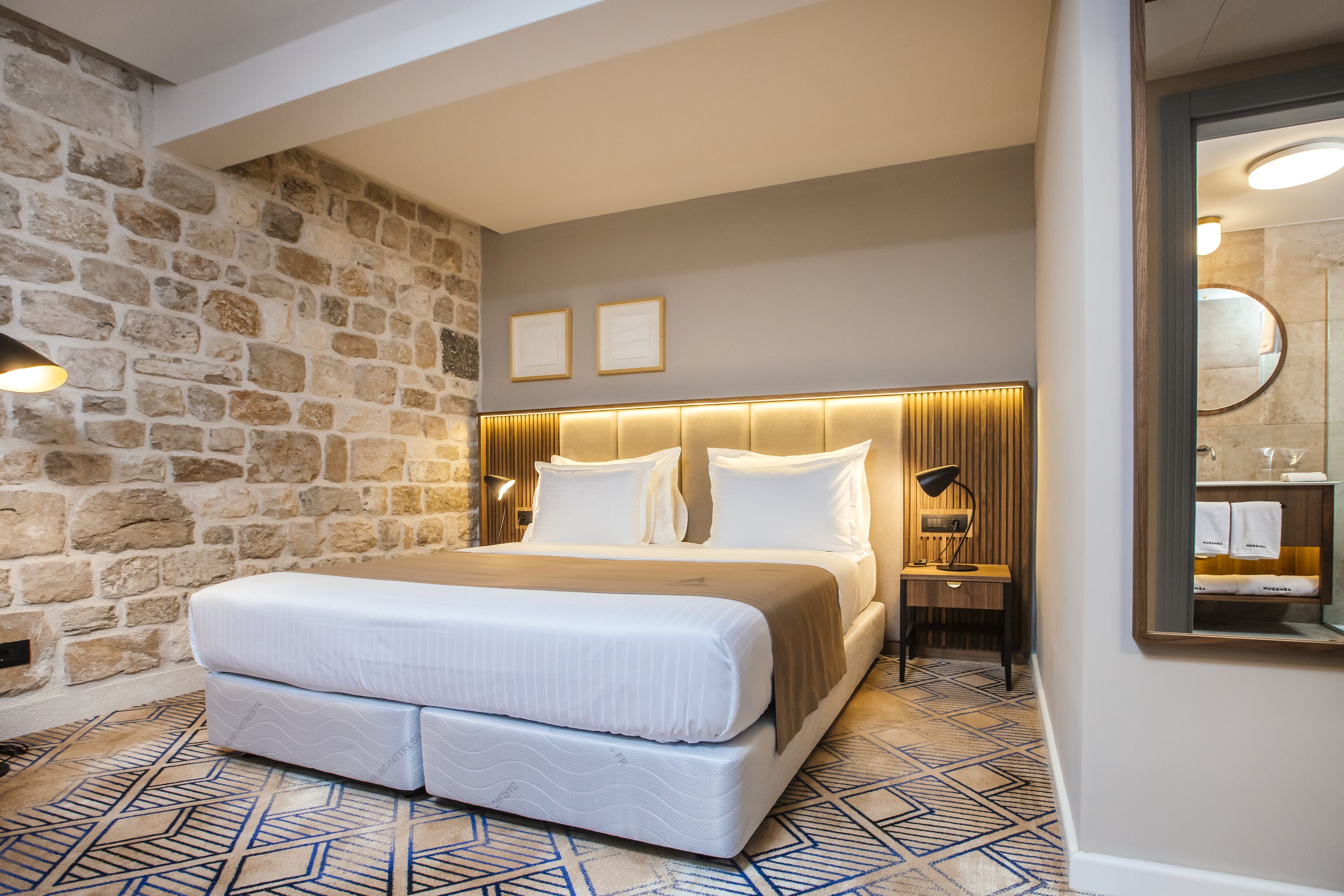 A hotel room with a stone wall, a large bed with a headboard, a nightstand, a lamp, and a mirror.