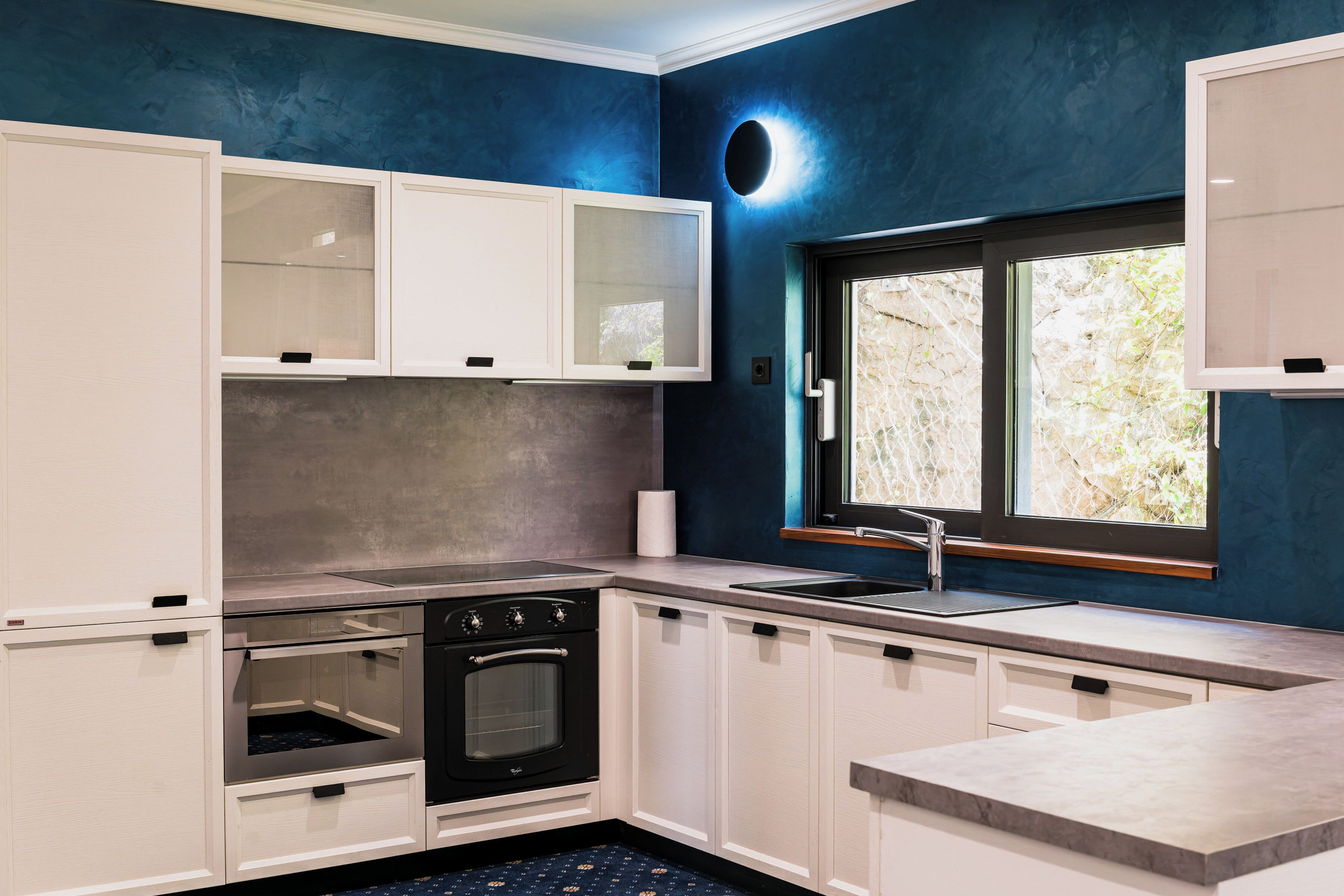 Een moderne keuken met witte kasten, een inbouwoven, een spoelbak en een raam met uitzicht op groen.