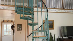 Uma sala de estar ampla com uma escada espiral turquesa, uma porta de madeira e uma televisão sobre um aparador de madeira.