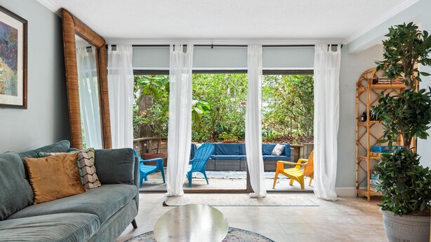 A living room with a grey sofa, a small round table, and a colorful rug. There are white curtains, a large window, and a view of a garden with chairs and a table outside. - Spacious Family or Couples Condo - Walk to Coligny Beach & Shops (Hilton Head Island)