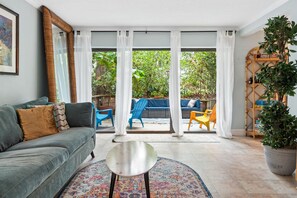 A living room with a grey sofa, a small round table, and a colorful rug. There are white curtains, a large window, and a view of a garden with chairs and a table outside.