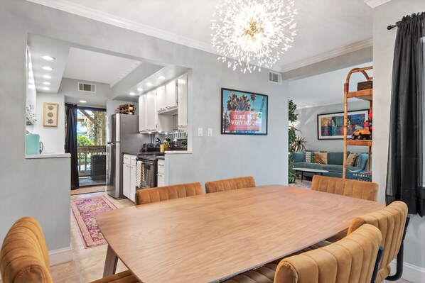 A modern dining area with a wooden table and chairs, a kitchen in the background, and a large chandelier.
