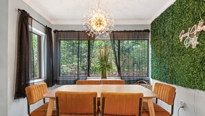 A dining area with a round table, six chairs, a chandelier, and a view of trees through large windows. - Spacious Family or Couples Condo - Walk to Coligny Beach & Shops (Hilton Head Island)