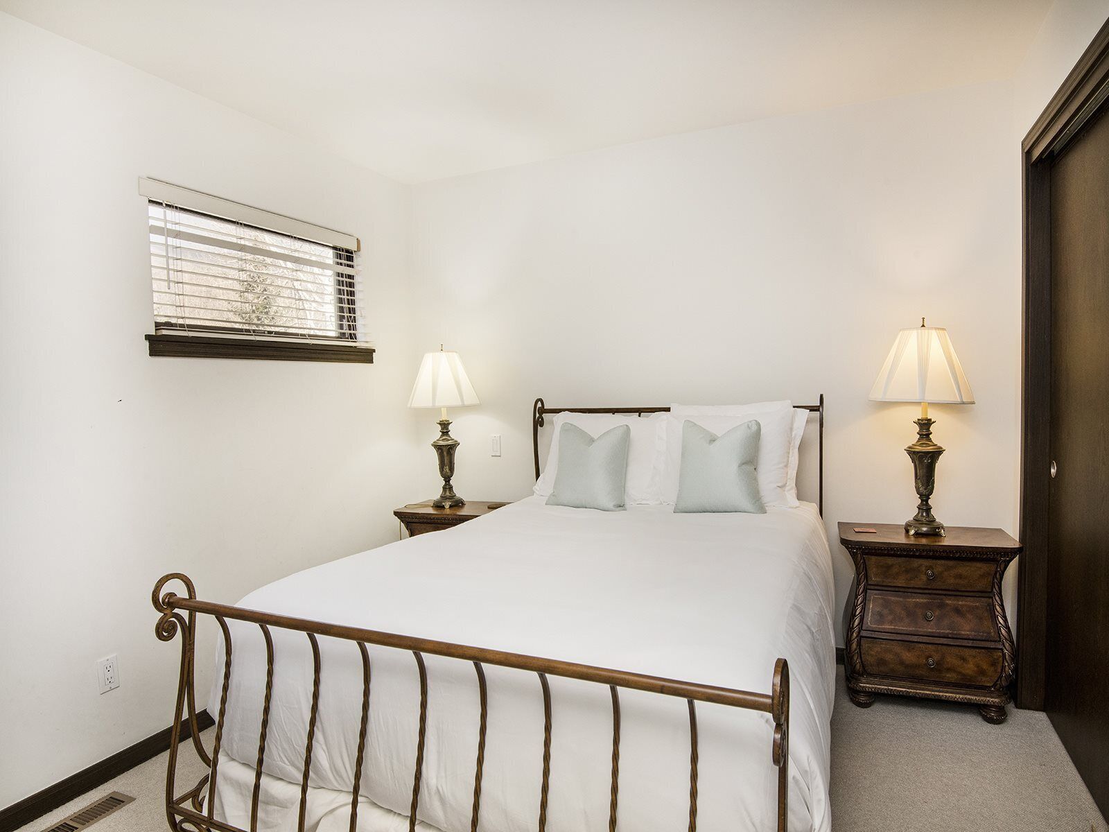 A bedroom with a metal bed frame, two bedside tables with lamps, a window with blinds, and a dark wooden door.