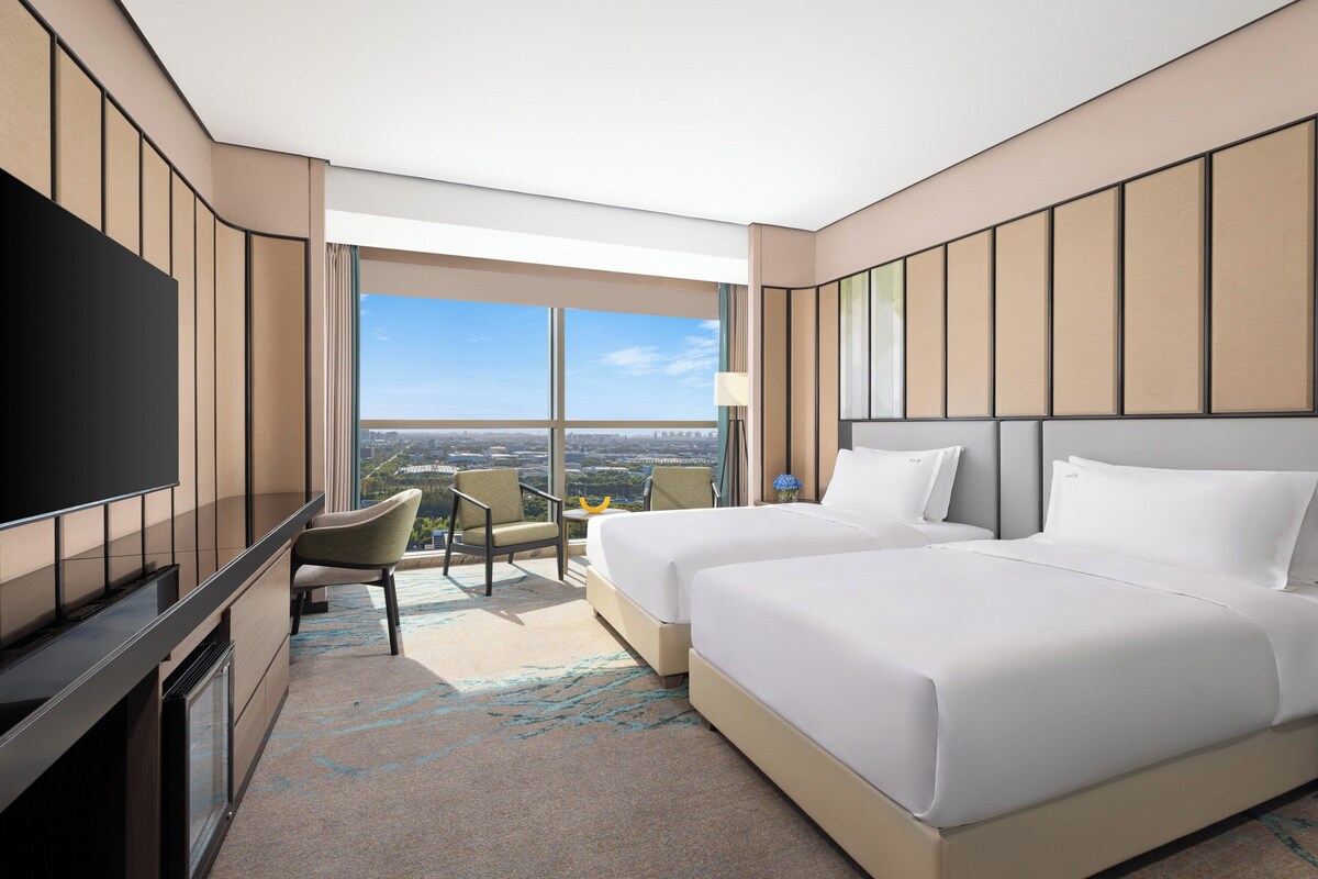 A hotel room with two beds, a TV, a desk, and a view of the city.