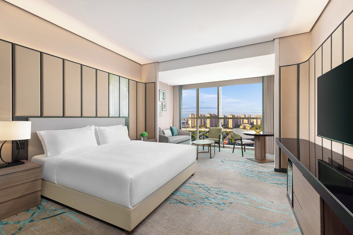 A modern hotel room with a large bed, a desk, a chair, a TV, and a view of the city.