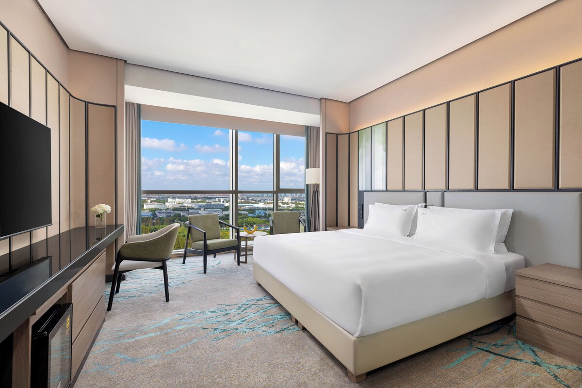 A modern hotel room with a large bed, a flat-screen TV, a desk, and a chair. The room has large windows offering a city view.