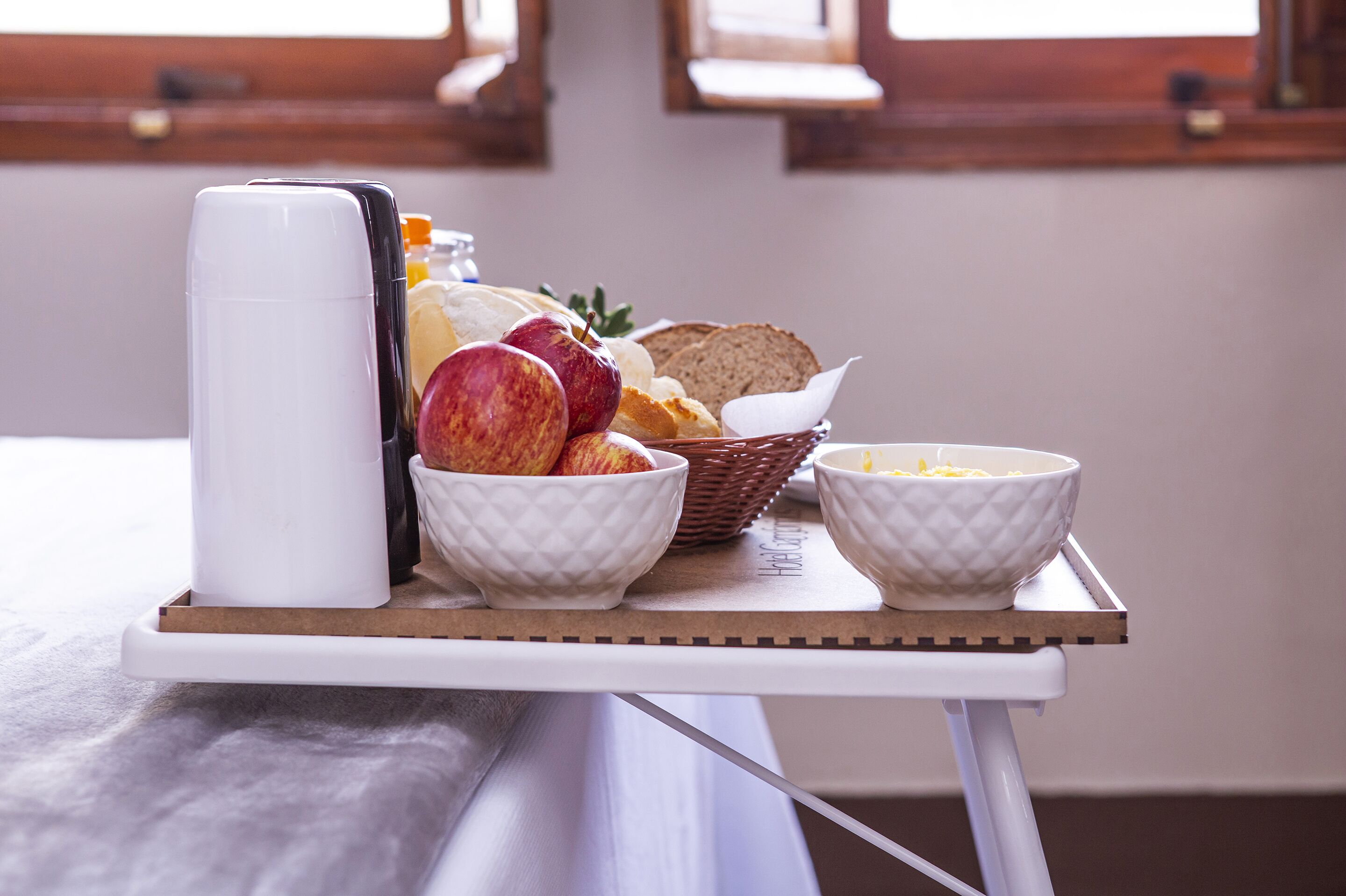 A breakfast tray with a coffee maker, bowls of cereal, apples, bread, and a basket of bread on a table.