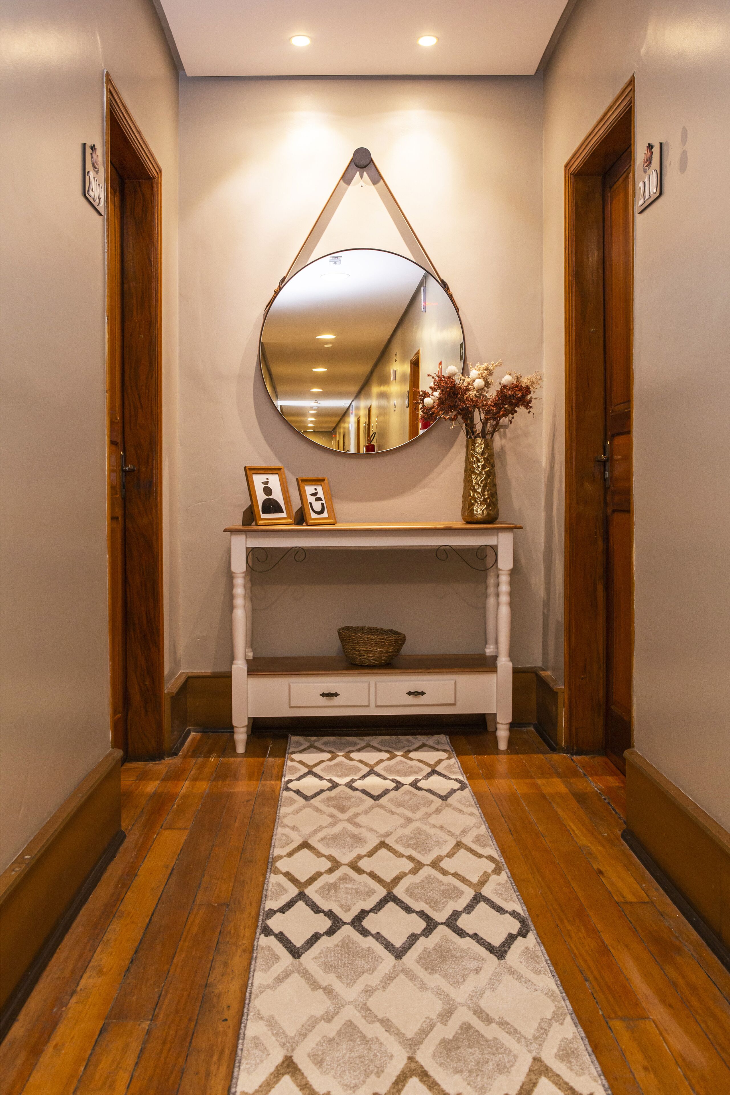 A hallway with a wooden floor, a patterned rug, a white console table with a mirror, a vase with flowers, and framed pictures.