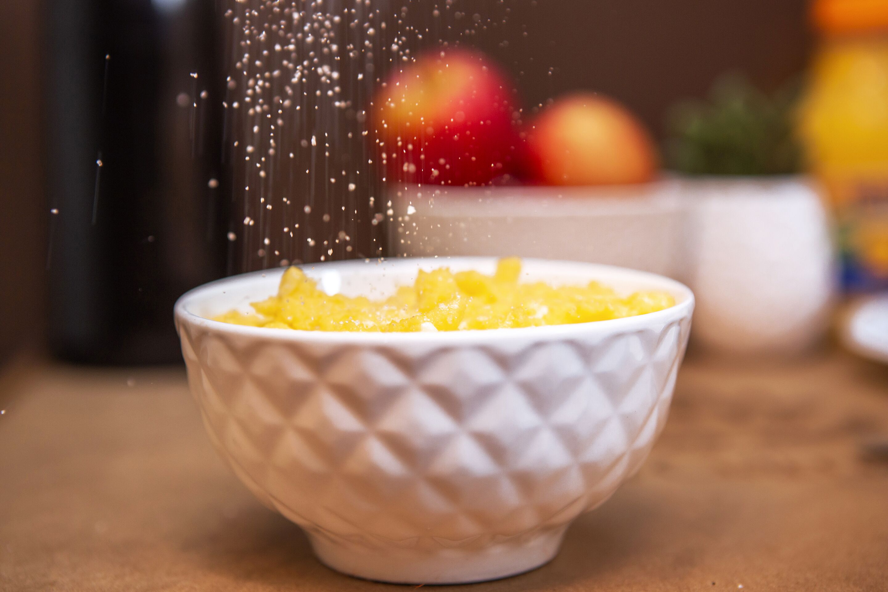 A white bowl with a textured pattern, containing a yellow substance, possibly food, with particles being sprinkled over it.