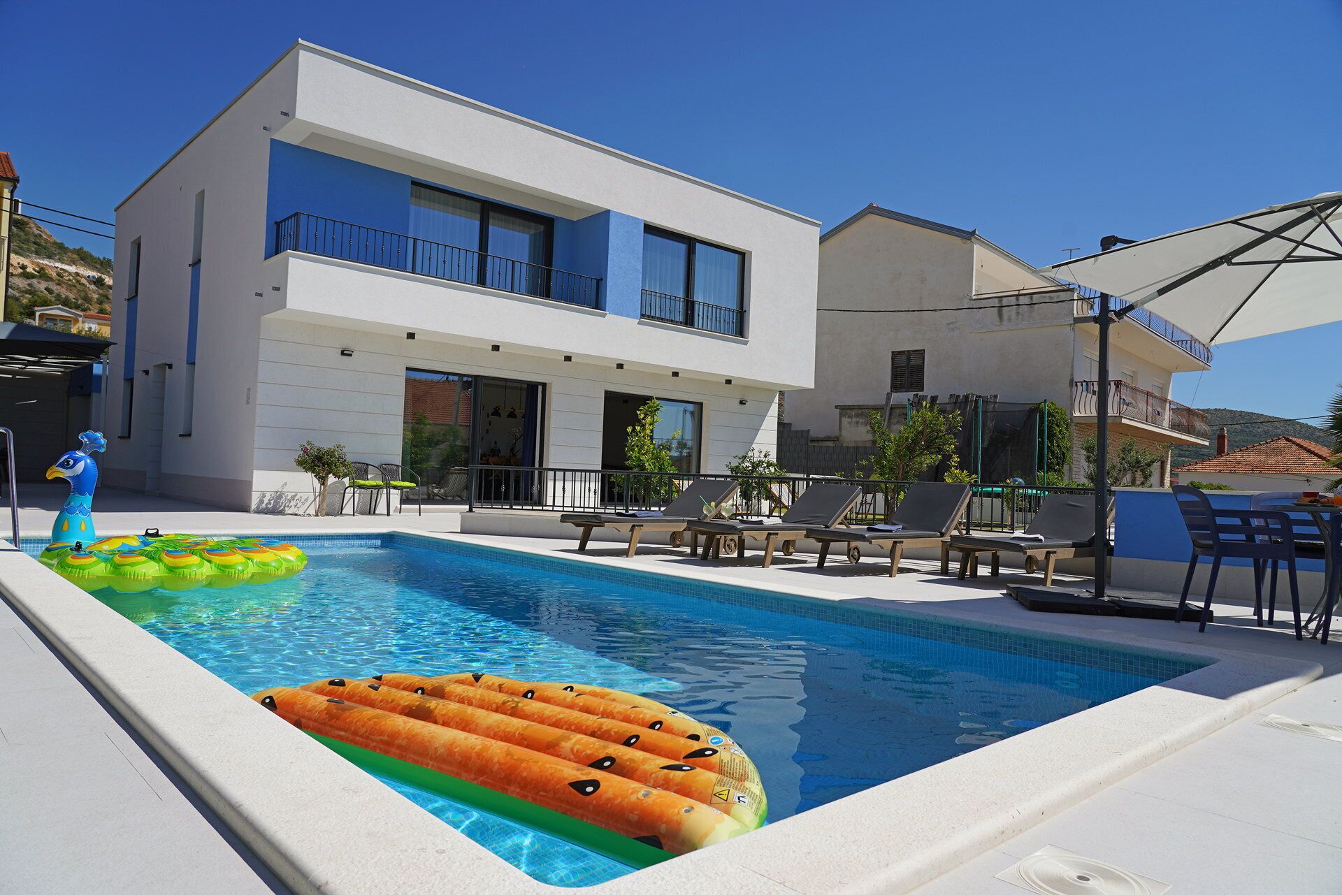 A modern two-story building with a swimming pool, lounge chairs, and a sun umbrella.