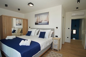 Une chambre moderne avec un grand lit, une armoire, un tableau représentant des voiliers et une salle de bain visible à travers une porte ouverte.