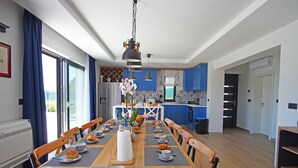 Een moderne keuken met blauwe kastjes, een eethoek met een houten tafel gedekt voor het ontbijt, en een groot raam met gordijnen.