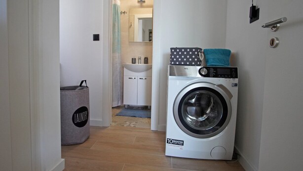 A laundry room with a front-loading washing machine, a grey laundry basket, and a bathroom visible in the background. - Villa Lady Blue with Heated Pool (Seget Donji)