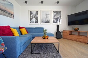 Un salon avec un canapé bleu, des coussins colorés, une table basse sur laquelle on a posé des fruits, un téléviseur et des tableaux encadrés accrochés au mur.