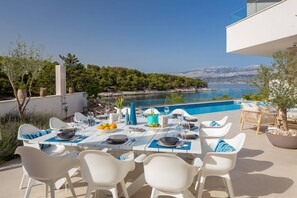 Un área de comedor al aire libre con vistas a un lago y montañas, sillas blancas y una mesa preparada para dos.