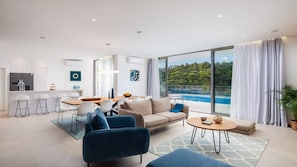 A modern living room with a sofa, chairs, a dining table, and a view of a pool and greenery.