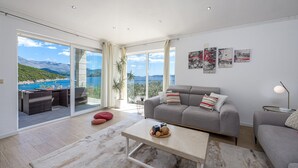 A spacious living room with a grey sofa, a coffee table, and a view of a mountainous coastline through large sliding glass doors. - Villa Auras with Heated Pool (Slano)