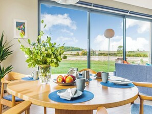 Een eetgedeelte met een tafel voor vier personen, een vaas met groene planten, een schaal met appels en uitzicht op een grasrijk landschap door grote ramen.