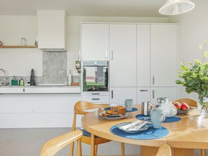 Moderni keittiö, jossa on valkoiset kaapit, sisäänrakennettu uuni sekä ruokailutila, jossa on puinen pöytä ja tuolit.