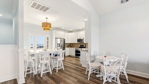 A modern kitchen with white cabinetry, a central island, and a dining area with white chairs. - Papa’s Porch, 8BR Beach House, Private Pool, Elevator, Sleeps 20 (Ocean Isle Beach)