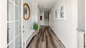 A hallway with white walls, a wooden floor, and a bench. There are framed pictures on the walls and a mirror on the left. - Papa’s Porch, 8BR Beach House, Private Pool, Elevator, Sleeps 20 (Ocean Isle Beach)