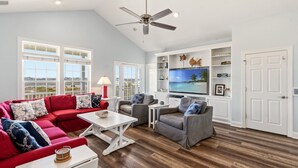 A spacious living room with a red sectional sofa, grey armchairs, a flat-screen TV, and a ceiling fan. - Papa’s Porch, 8BR Beach House, Private Pool, Elevator, Sleeps 20 (Ocean Isle Beach)