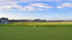 A golf course with a flagstick in the foreground, buildings in the background, and a clear sky.