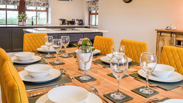 A dining area with yellow upholstered chairs, a wooden table set with white plates, bowls, and wine glasses. - 4 bedroom accommodation in Carlisle (Carlisle)