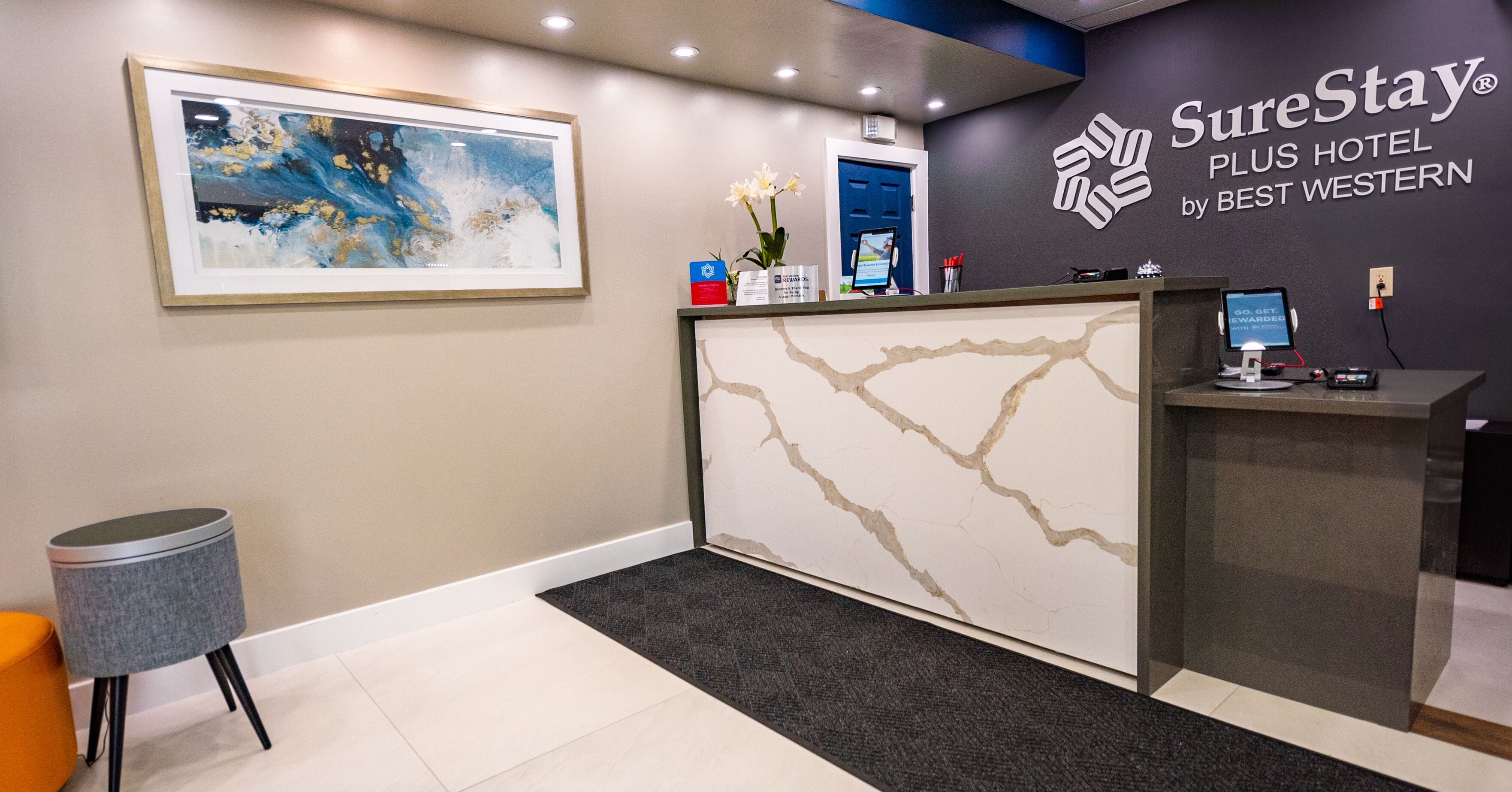 Zona de recepción de un hotel con un mostrador de mármol, un cuadro abstracto enmarcado, una puerta azul y un letrero de SureStay Plus Hotel.
