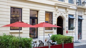 An outdoor cafe with red umbrellas, tables, and chairs, and a building with large windows and a door. - Hotel De Cambis (Avignon)
