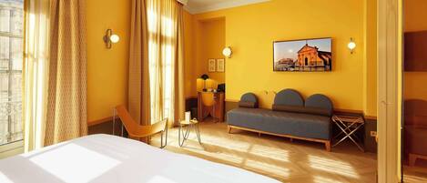 A hotel room with a large bed, a sofa, a desk, and a chair. There is a window with curtains, a painting on the wall, and a lamp.