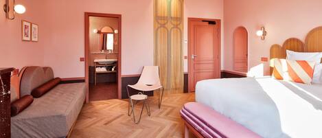 A hotel room with a wooden herringbone floor, a sofa, a bed with a pink mattress, a bench, a chair, a wardrobe, and a mirror.