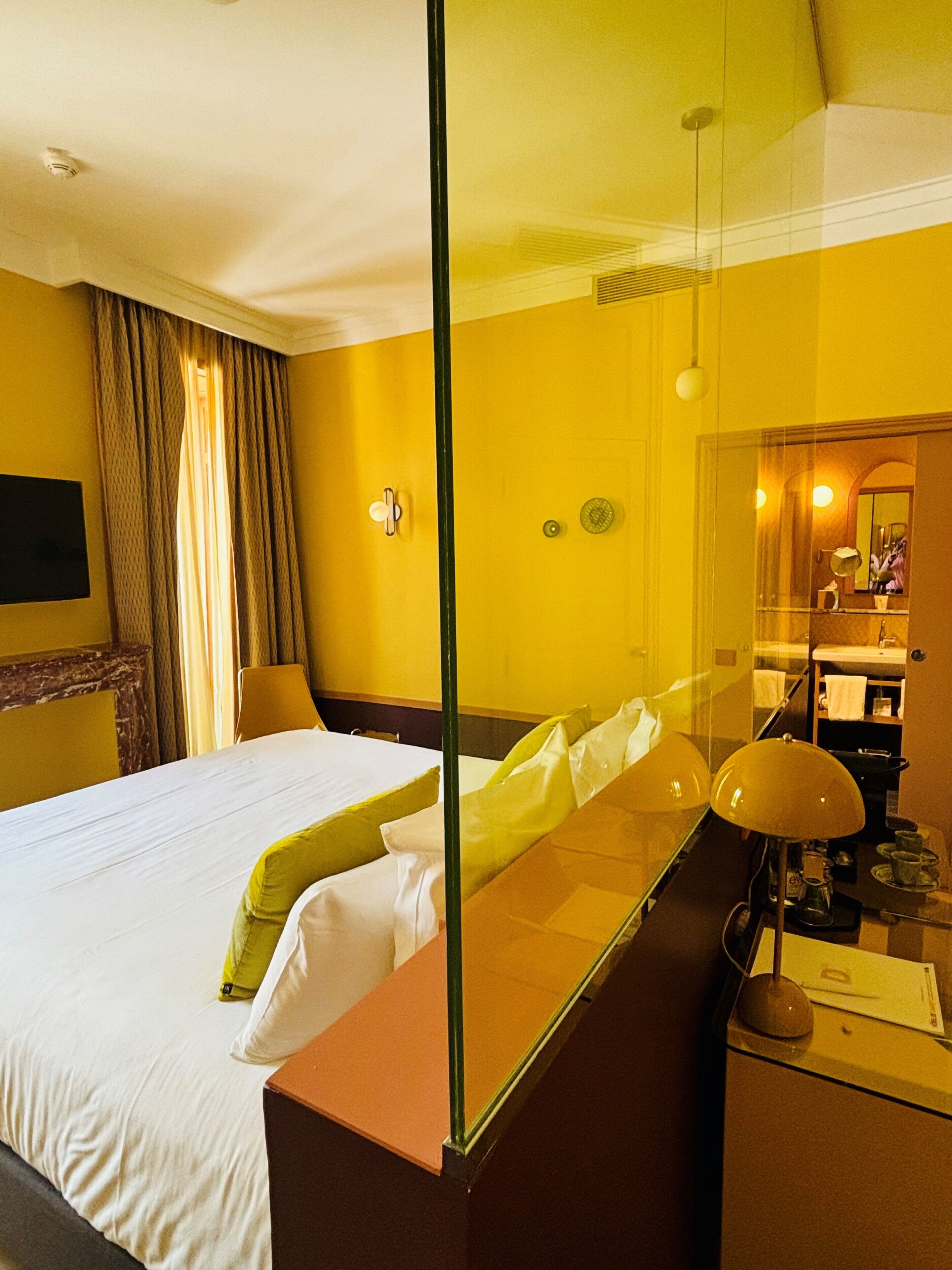 Une chambre d’hôtel équipée d’un lit, d’un bureau, d’une lampe, d’un miroir et d’une salle de bain visible à travers une paroi en verre.