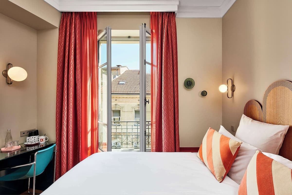 A hotel room with a bed, a desk with a phone and a coffee maker, red curtains, and a view of buildings through a glass door.