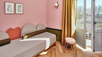 A room with a bed, a small stool, a wall-mounted light, and a window with a view of buildings.