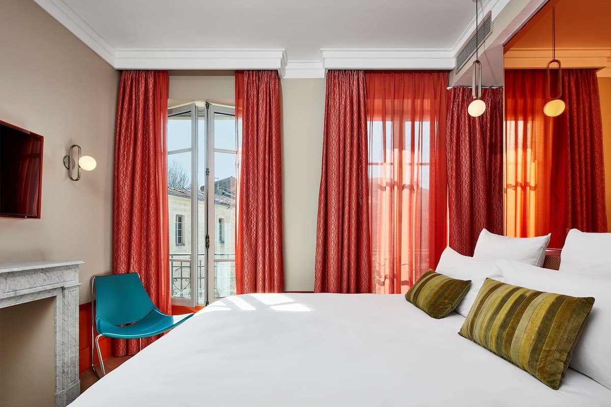 A hotel room with a large bed, red curtains, a blue chair, and a fireplace.