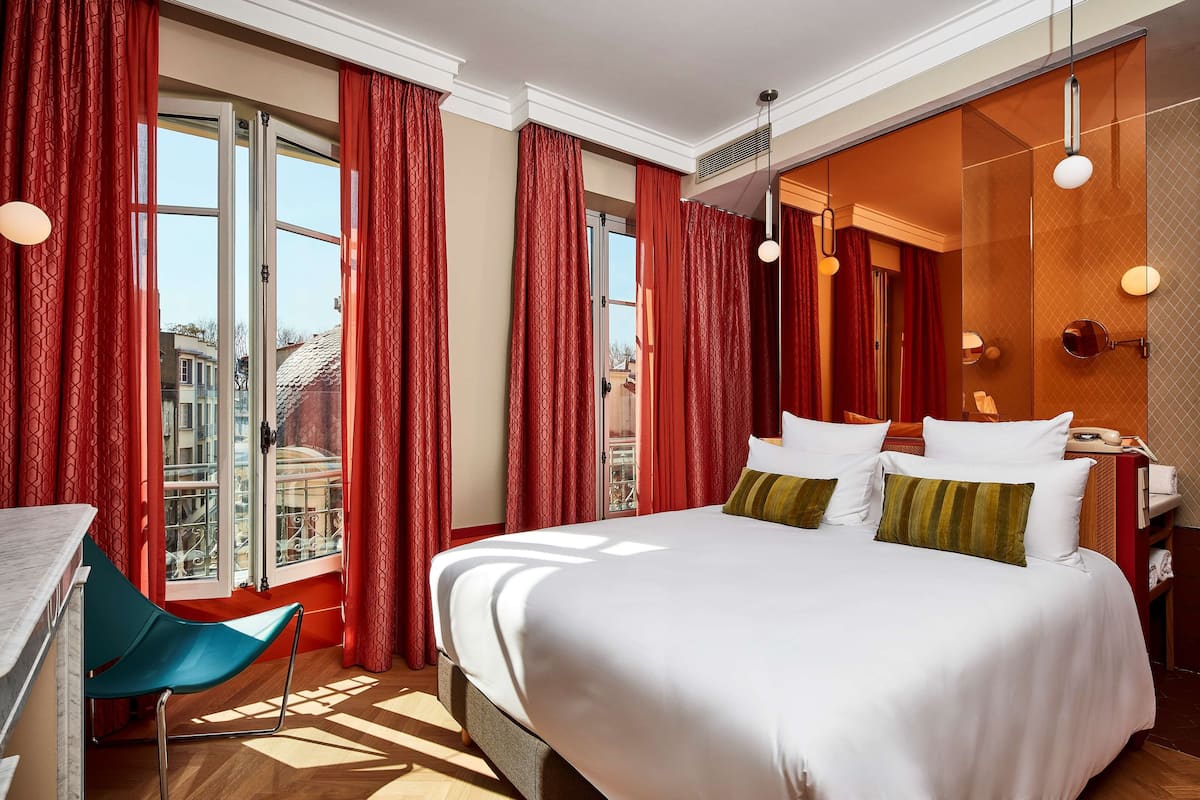 A hotel room with a large bed, red curtains, a desk, and a view of buildings outside.