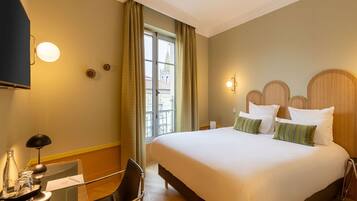 A hotel room with a large bed, a desk with a chair, a bottle, glasses, and a tray.