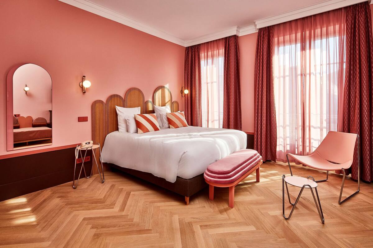 A bedroom with a large bed, a chair, a small stool, a mirror, and a window with curtains.